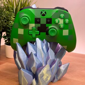 May include: A green and black video game controller with a Minecraft design is displayed on a blue and purple crystal-shaped stand.