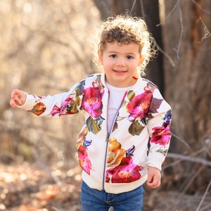May include: A young child with curly hair wearing a white floral bomber jacket and blue jeans. The child is standing in a wooded area with branches and leaves in the background.
