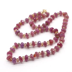 May include: A necklace featuring a strand of pink and purple gemstones, with smaller clear and yellow beads. The necklace, with a gold clasp, is displayed against a white backdrop. The gemstones are of varying sizes.