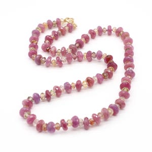 May include: A necklace with pink and yellow gemstones. The necklace has a gold clasp and is made up of various shapes and sizes of polished stones. The gemstones are a mix of pink and yellow hues, creating a colourful and elegant accessory.