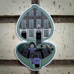 May include: A green heart-shaped locket with a miniature house with windows and doors. The locket is open, revealing a miniature scene with a figure in a purple robe standing in a room with a black and white patterned rug. The house has the numbers 11 and 12 on the doors.