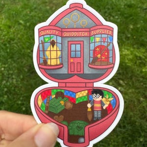 May include: A die-cut sticker shaped like a heart-shaped compact case featuring a red storefront labelled Quality Quidditch Supplies on the top half and an interior scene with a character holding a broomstick on the bottom half.