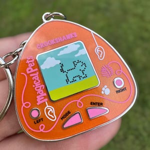 May include: Orange keychain featuring a retro digital pet game. The screen displays a pixelated dog under a blue sky. The keychain has pink buttons and the text "Magical Pets" and "Crookshanks". A silver keyring is attached.