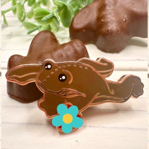 May include: A gold-tone enamel pin of a brown frog holding a blue flower with a yellow center. The frog is jumping and has a white eye with a black pupil.