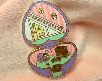 Puede incluir: Un pin de esmalte de color dorado con forma de medallón en forma de corazón. El medallón está abierto, revelando un interior en miniatura con una pared a rayas rosa y blanca, una escalera, una chimenea y una figura de pie en la habitación.