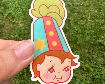 May include: A sticker featuring a vintage-style illustration of a child's face wearing a party hat. The hat is teal, red, and yellow, with a star and three circles. The child has rosy cheeks, brown hair, and a slightly grumpy expression.