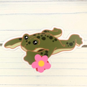 May include: A green frog with brown spots holding a pink flower with a yellow center. The frog is swimming with its legs outstretched.