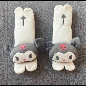 May include: A pair of white plush car seat headrests with black and gray Hello Kitty characters. The characters have pink cheeks and a red skull and crossbones on their foreheads. The headrests have an arrow pointing upwards.