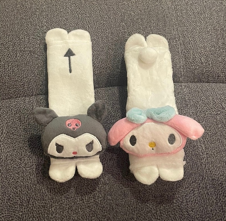 May include: Two plush car seat headrests, one with a black and white skull design and the other with a pink and blue Hello Kitty design.