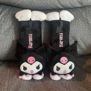 May include: A pair of black plush car seat headrests featuring a cartoon character with pink accents. The character has a skull on its forehead and the text "Kuromi" is embroidered on each headrest.