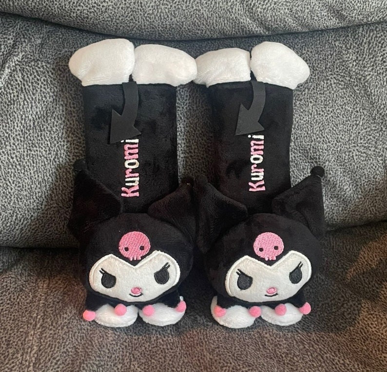 May include: A pair of black plush car seat headrests featuring a cartoon character with pink accents. The character has a skull on its forehead and the text "Kuromi" is embroidered on each headrest.
