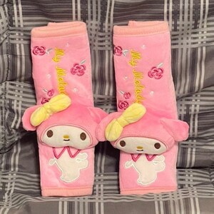 May include: Two pink car seat headrest covers with embroidered My Melody characters and roses. The characters are wearing yellow bows and have white faces with black eyes and mouths.