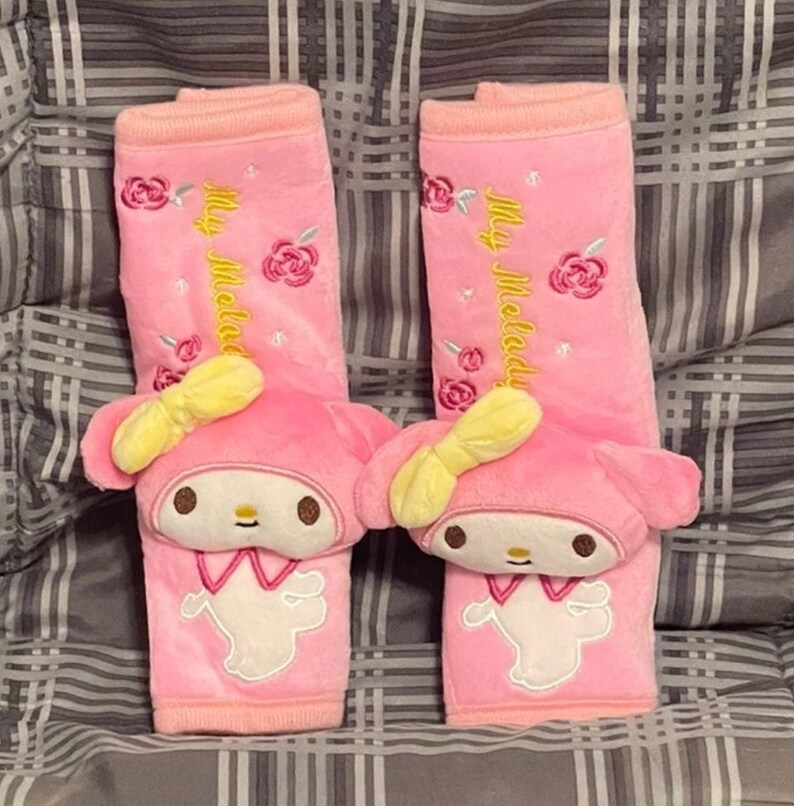 May include: Two pink car seat headrest covers with embroidered My Melody characters and roses. The characters are wearing yellow bows and have white faces with black eyes and mouths.