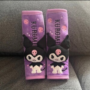 May include: Two purple plush car seat headrests with a black and white Kuromi character. The headrests have pink skulls and glitter accents. The text "Kuromi" is embroidered on each headrest.