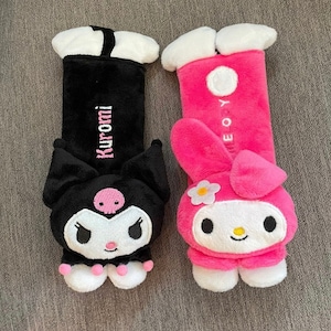 May include: Two plush car seat headrests featuring popular Japanese characters. The black headrest features a black cat with pink accents and the text "Kuromi" embroidered on the back. The pink headrest features a pink rabbit with white accents and the text "My Melody" embroidered on the back.
