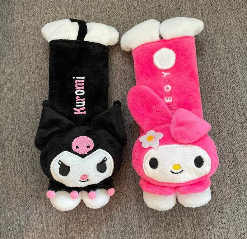 May include: Two plush car seat headrests featuring popular Japanese characters. The black headrest features a black cat with pink accents and the text "Kuromi" embroidered on the back. The pink headrest features a pink rabbit with white accents and the text "My Melody" embroidered on the back.