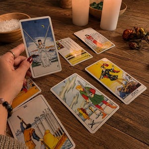 Pu&ograve; includere: Una collezione di carte dei tarocchi sparse su una superficie in legno, tra cui il Due di Spade, il Cinque di Spade, il Sei di Spade e una carta Joker. Le carte presentano illustrazioni dettagliate con figure in varie pose, sullo sfondo di due candele accese.