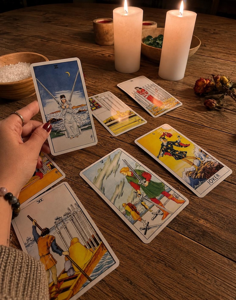 Puede incluir: Una colecci&oacute;n de cartas de tarot extendidas sobre una superficie de madera, incluyendo el Dos de Espadas, el Cinco de Espadas, el Seis de Espadas y una carta de Joker. Las cartas presentan ilustraciones detalladas con figuras en varias poses, junto a dos velas encendidas.