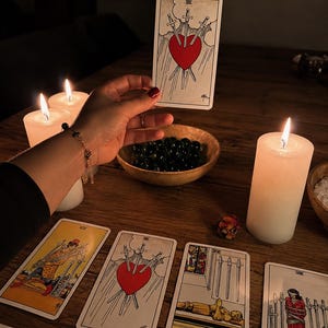Op de afbeelding: Een tarotkaart met een groot rood hart doorboord door drie zilveren zwaarden tegen een bewolkte lucht, vastgehouden boven een houten tafel met brandende witte stompkaarsen, een houten kom met donkere stenen en diverse andere tarotkaarten.