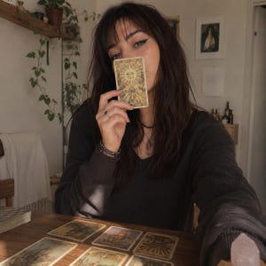 Op de afbeelding: Een rechthoekige tarotkaart met een centrale zonne-illustratie met een gezicht, omringd door stralen en bloemmotieven, voor het gezicht gehouden. De kaart is beige met gouden en bruine inktgegevens en een klassiek hemels ontwerp.
