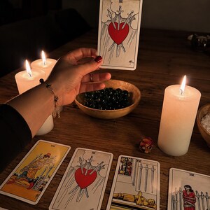 Puede incluir: Una carta de tarot que muestra un coraz&oacute;n rojo atravesado por tres espadas sobre un cielo nublado, sostenida sobre una mesa de madera con velas blancas encendidas, un cuenco de madera con piedras oscuras y otras cartas de tarot.