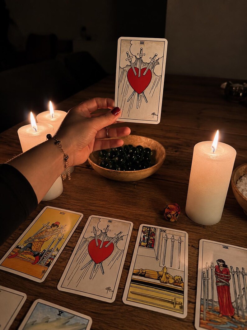Puede incluir: Una carta de tarot que muestra un coraz&oacute;n rojo atravesado por tres espadas sobre un cielo nublado, sostenida sobre una mesa de madera con velas blancas encendidas, un cuenco de madera con piedras oscuras y otras cartas de tarot.
