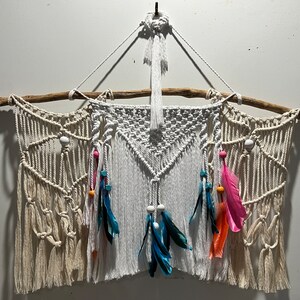 May include: A white macrame wall hanging with a driftwood branch at the top. The hanging features a geometric pattern with white beads and colourful feathers.