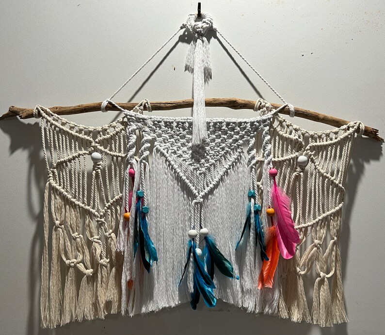 May include: A white macrame wall hanging with a driftwood branch at the top. The hanging features a geometric pattern with white beads and colourful feathers.