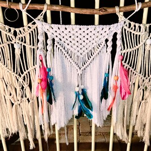 May include: A white macrame wall hanging with a triangular design and fringe. The hanging is decorated with white beads, pink, blue, and orange feathers.