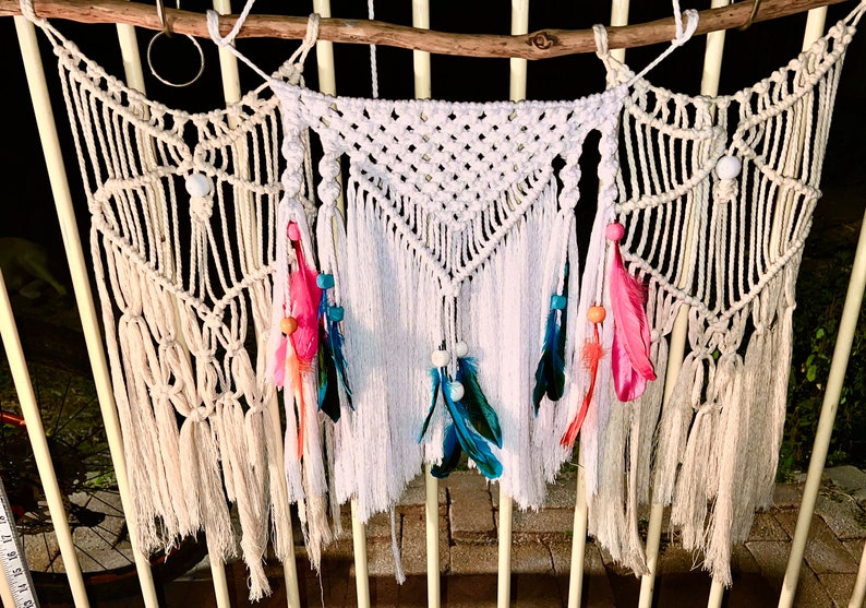 May include: A white macrame wall hanging with a triangular design and fringe. The hanging is decorated with white beads, pink, blue, and orange feathers.