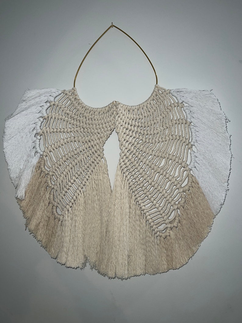 Pu&ograve; includere: Un arazzo da parete in macram&egrave; beige e bianco con un anello in metallo dorato nella parte superiore. L'arazzo ha un bordo a frangia e un motivo geometrico.