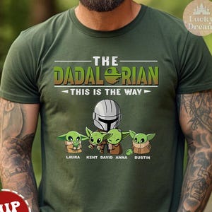 May include: Green t-shirt with a Star Wars themed design featuring a Mandalorian and four Baby Yoda characters. The text reads "The Dadalorian This is the Way" with the names Laura, Kent, David, Anna, and Dustin below the characters.