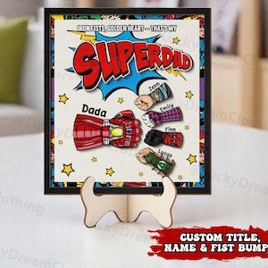May include: A black framed print with a comic book style design featuring the text "IRON FISTS, GOLDEN HEART - THAT'S MY SUPERDAD". The print features a cartoon image of a superhero's fist with the words "Dada", "Zenn", "Emily", "Finn", and "Lucas" written on the fists.