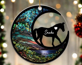 Könnte beinhalten: Ein Weihnachtsbaumschmuck im Stil von Buntglas mit einer schwarzen Silhouette eines Pferdes mit dem Namen "Smoke" an der Seite, das auf einem Halbmond mit einem Herzen über seinem Kopf steht. Der Hintergrund ist ein buntes Buntglasdesign einer Landschaft mit Bäumen, Bergen und einem See.