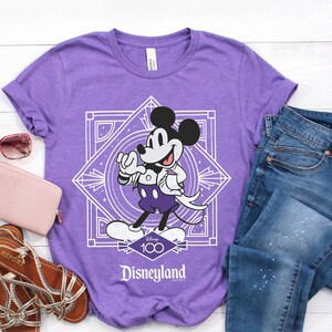 May include: A purple t-shirt featuring a graphic of Mickey Mouse in a tuxedo, with a white geometric design and the text "Disneyland Resort". The shirt is paired with blue jeans, sandals, a pink wallet, and sunglasses.
