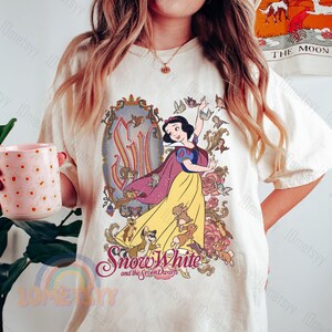 May include: A white t-shirt featuring a vintage-style illustration of Snow White and the Seven Dwarfs. The illustration depicts Snow White in a yellow dress with a red cape, surrounded by the dwarfs and forest animals. The text "Snow White and the Seven Dwarfs" is printed beneath the illustration.