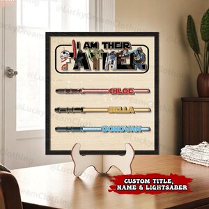 K&ouml;nnte beinhalten: Ein gerahmter Star Wars-Print mit dem Text "I AM THEIR FATHER" und drei Lichtschwert-Designs. Jedes Lichtschwert hat einen Namen: Chloe, Bella und Donovan. Der Druck wird auf einem Holzst&auml;nder pr&auml;sentiert.