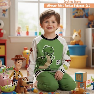 Puede incluir: Un niño lleva un conjunto de pijama raglán verde y blanco con un diseño de dinosaurio de dibujos animados. El conjunto de pijama incluye una camiseta de manga larga y pantalones a juego. El texto "All-over Print Raglan Pajamas Set" es visible.