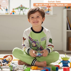 May include: A child wearing a green and white Buzz Lightyear pajama set. The long-sleeved top features Buzz Lightyear and the word "BUZZ". The pajama set includes matching green pants. The text "Custom Name All-over Print Raglan Pajamas Set" is visible.