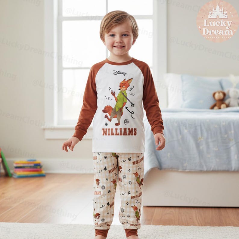 Peut inclure: Un enfant porte un pyjama Disney. Le haut est blanc avec des manches marron et pr&eacute;sente un dessin de renard de dessin anim&eacute; et le nom "WILLIAMS". Le pantalon de pyjama est beige avec un motif r&eacute;p&eacute;t&eacute; de renard et de texte. Le logo Disney est visible.