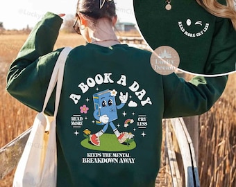 Pode incluir: Uma camisa de manga comprida verde com um personagem de livro de desenho animado nas costas. O personagem do livro está andando e tem as palavras "A Book A Day" acima dele. O texto "Read More Cry Less" está impresso na frente da camisa.
