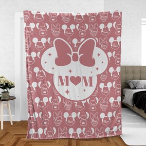 K&ouml;nnte beinhalten: Eine rosa Fleece-Decke mit einer wei&szlig;en Umrandung eines Mickey-Maus-Kopfes mit Schleife und dem Wort "MOM" in der Mitte. Der Hintergrund ist ein sich wiederholendes Muster von Mickey-Maus-K&ouml;pfen mit den Namen "Jake", "Bree" und "Sam" darauf.