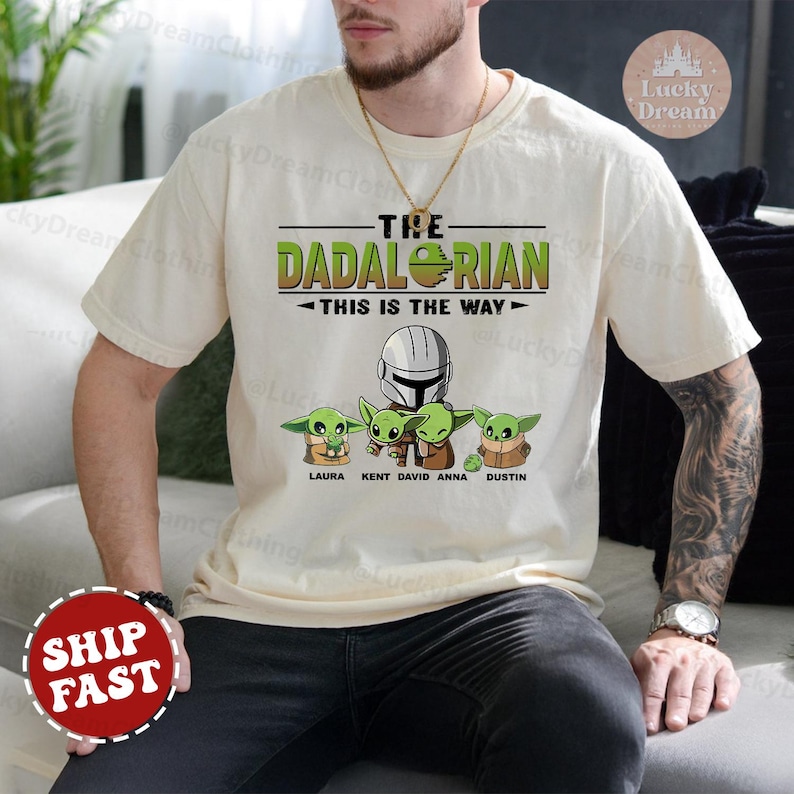 May include: A beige t-shirt with a green, white, and black graphic of a Star Wars character with the text "The Dadalorian - This is the Way" and five smaller green characters with names below them.