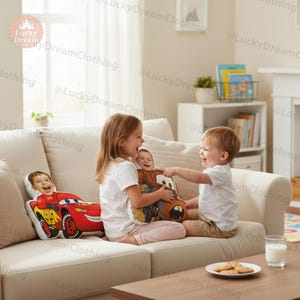 May include: Two custom-printed throw pillows featuring children's faces and cartoon car designs. One pillow showcases a red race car with the number 95, while the other depicts a brown tow truck. The pillows are on a cream-colored sofa.