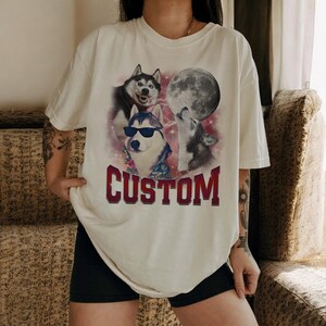 May include: A white t-shirt with a graphic of three husky dogs howling at the moon. The text "CUSTOM" is printed in red below the dogs.