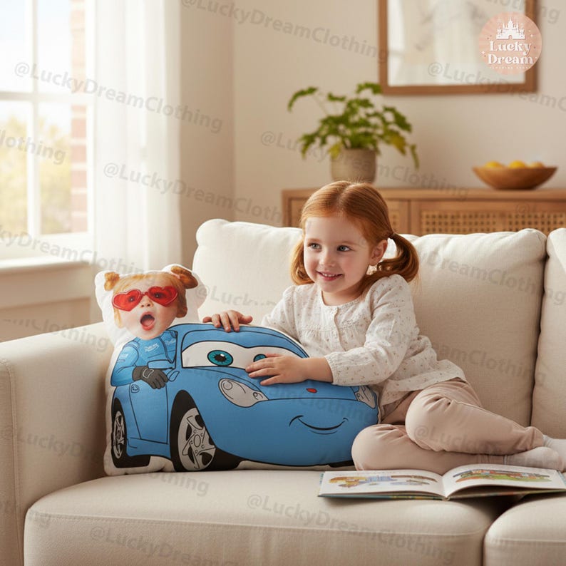 May include: A blue car-shaped pillow with a cartoon image of a child wearing heart-shaped sunglasses and a racing suit. The pillow is on a white couch. A young girl with red hair is sitting on the couch, holding the pillow.