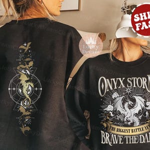K&ouml;nnte beinhalten: Schwarzer Pullover mit einem goldenen Drachen-Design auf der R&uuml;ckseite und dem Text "ONYX STORM THE BIGGEST BATTLE YET BRAVE THE DARK" auf der Vorderseite.