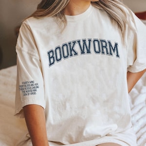 Pode incluir: Uma camiseta branca com o texto "BOOKWORM" impresso em azul marinho, em uma fonte de estilo universit&aacute;rio. O texto &eacute; arqueado na parte frontal da camiseta. A camiseta tem um aspecto desgastado e vintage. O texto "FOURTH WING FROM BLOOD AND ASH THEIR VICIOUS DARLING THE NERVER KING DEN OF VIPERS" est&aacute; impresso na manga esquerda da camiseta.