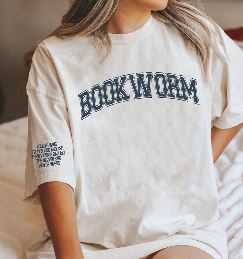 Pode incluir: Uma camiseta branca com o texto "BOOKWORM" impresso em azul marinho, em uma fonte de estilo universit&aacute;rio. O texto &eacute; arqueado na parte frontal da camiseta. A camiseta tem um aspecto desgastado e vintage. O texto "FOURTH WING FROM BLOOD AND ASH THEIR VICIOUS DARLING THE NERVER KING DEN OF VIPERS" est&aacute; impresso na manga esquerda da camiseta.