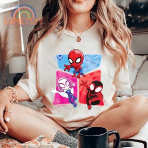 May include: A white t-shirt with a graphic of three cartoon Spider-Man characters. The characters are in different poses and colors, including red, blue, black, and white.